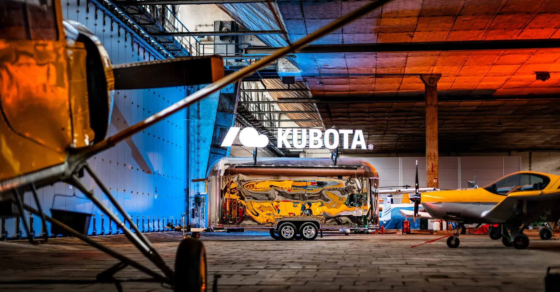 Zdjęcie przedstawiające lustrzaną przyczepę eventową SHINE z uniesioną przeszkloną ścianą boczną, zaparkowaną w nowoczesnym industrialnym wnętrzu z widokiem na miasto, działającą jako nowoczesny mobilny showroom.